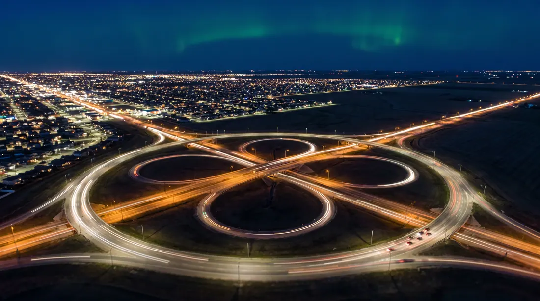 Night aerial of a ring-road interchange on the edge of a prairie city, light trails curving around the ramps