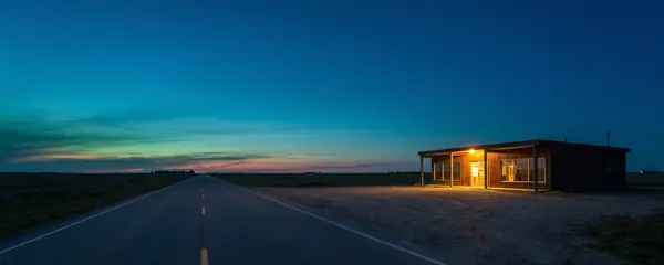 A two-lane prairie highway at blue hour with one low roadside building lit from inside